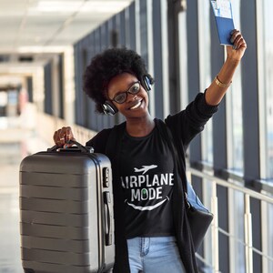 Może przedstawiać: Kobieta w czarnym t-shircie z napisem "Airplane Mode" i białym nadrukiem samolotu. Trzyma czarną walizkę i paszport. Uśmiecha się i patrzy w obiektyw.