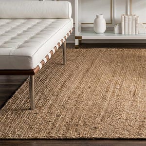 May include: A rectangular area rug made of natural tan woven jute fibres with a coarse, textured surface. The rug is positioned on a dark wooden floor, extending from the foreground towards a white console table, a white daybed, and a white armchair.