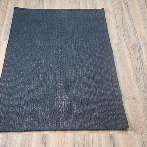 May include: A dark gray rectangular rug with a textured woven pattern. The rug is lying flat on a light wood floor.