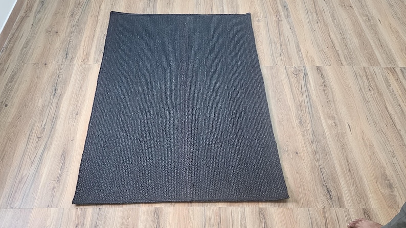 May include: A dark gray rectangular rug with a textured woven pattern. The rug is lying flat on a light wood floor.