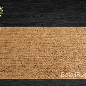 May include: A rectangular, natural-toned jute rug with a woven texture. The rug is placed on a dark wood surface, with a potted plant in the upper left corner. The rug's edges are neatly finished.