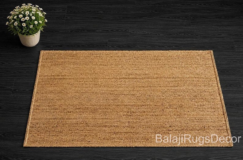 May include: A rectangular, natural-toned jute rug with a woven texture. The rug is placed on a dark wood surface, with a potted plant in the upper left corner. The rug's edges are neatly finished.