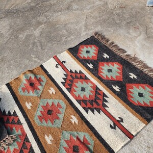 May include: A handwoven rug with a geometric pattern in shades of brown, red, white, and green. The rug has a fringe border and is made of natural fibers.