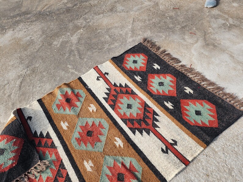 May include: A handwoven rug with a geometric pattern in shades of brown, red, white, and green. The rug has a fringe border and is made of natural fibers.