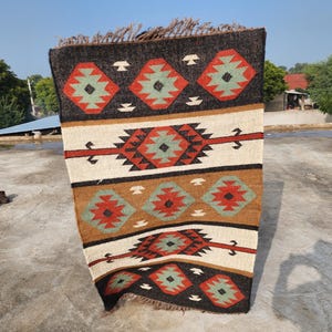 May include: A rectangular rug with a geometric pattern in shades of brown, red, white, and green. The rug has a fringe border.
