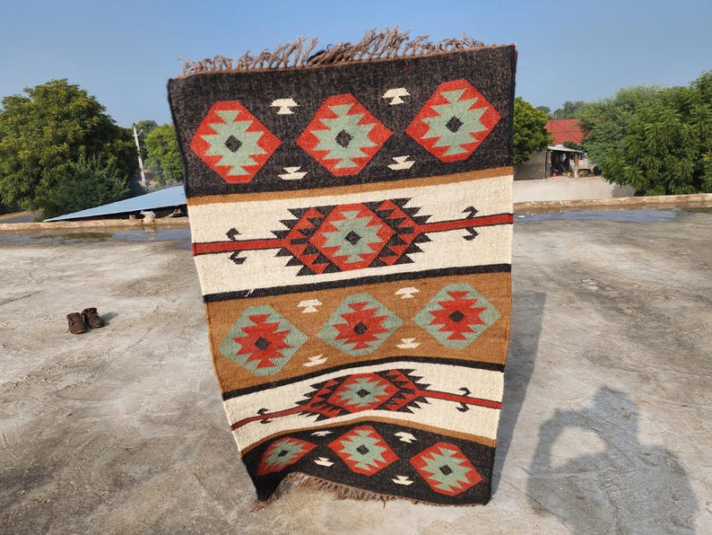 May include: A rectangular rug with a geometric pattern in shades of brown, red, white, and green. The rug has a fringe border.
