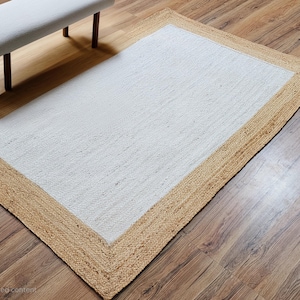 May include: Cream-coloured rectangular jute rug with a natural fibre border. The rug is placed on a wooden floor, with a white bench and dark legs in the background. The rug's design adds a touch of natural texture to the room.