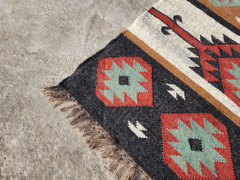 May include: A black, red, and green patterned rug with fringe. The rug has a geometric design with a white and brown background.