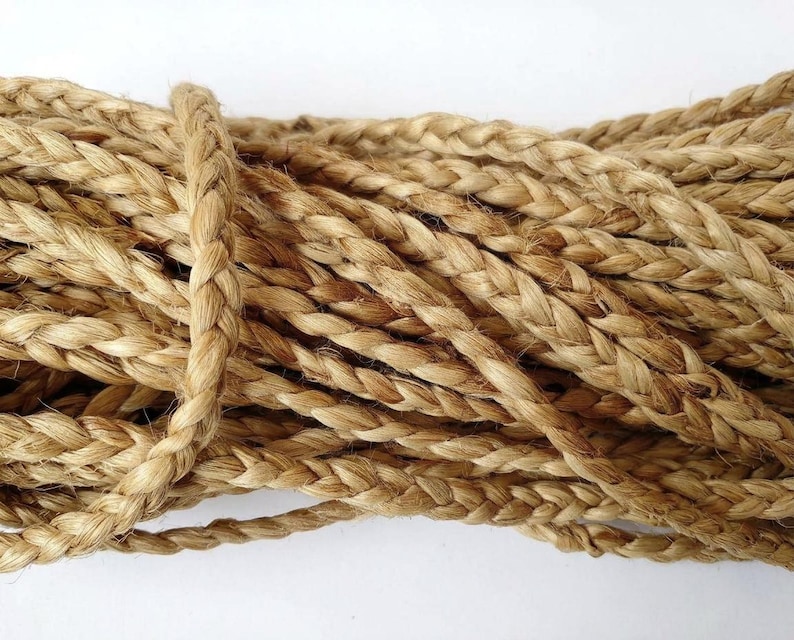 May include: A coiled length of natural tan jute rope featuring a tight, three-strand braided construction. The fibers exhibit a fibrous, textured appearance with varying shades of light brown and golden tan throughout the braided cord.