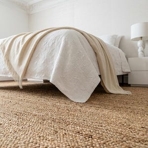 May include: A large, rectangular, natural-colored woven jute area rug with a thick, textured, braided fiber construction. The rug is positioned underneath a bed frame, covering the floor with its dense, organic, tan-colored material.