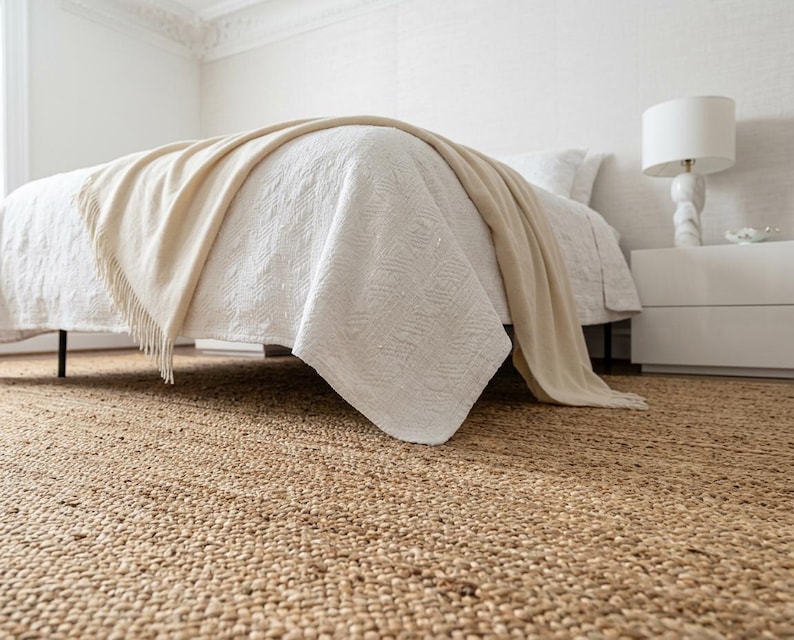 May include: A large, rectangular, natural-colored woven jute area rug with a thick, textured, braided fiber construction. The rug is positioned underneath a bed frame, covering the floor with its dense, organic, tan-colored material.