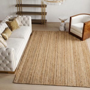 May include: A rectangular area rug crafted from natural woven jute fibers in a light tan color. The rug features a consistent, textured braided pattern throughout its surface, lying flat on a light-colored wooden floor in a living room setting.