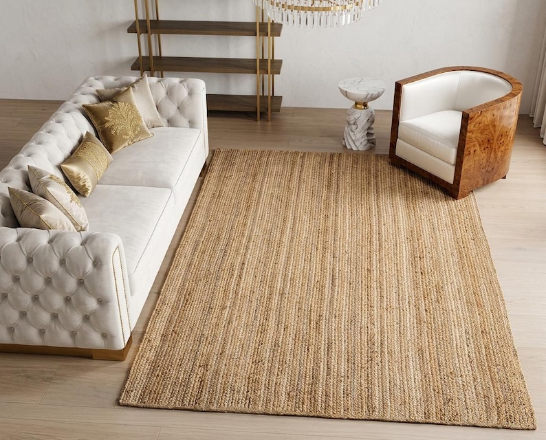May include: A rectangular area rug crafted from natural woven jute fibers in a light tan color. The rug features a consistent, textured braided pattern throughout its surface, lying flat on a light-colored wooden floor in a living room setting.