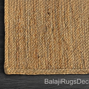 May include: Close-up of a rectangular jute rug with a braided border. The rug has a natural, light brown color and a woven texture. The text "BalajiRugsDecor" is visible at the bottom.