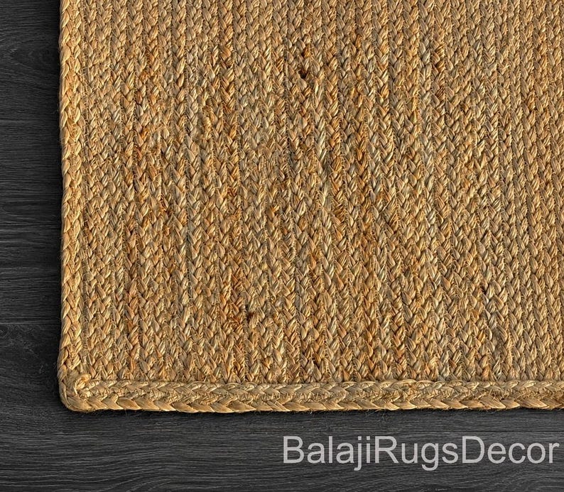 May include: Close-up of a rectangular jute rug with a braided border. The rug has a natural, light brown color and a woven texture. The text "BalajiRugsDecor" is visible at the bottom.