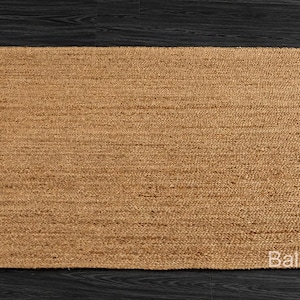 May include: A rectangular, woven jute rug in a natural tan color. The rug has a simple, textured design and is placed on a dark wood surface. The rug's edges are neatly finished, and the overall aesthetic is minimalist and earthy.