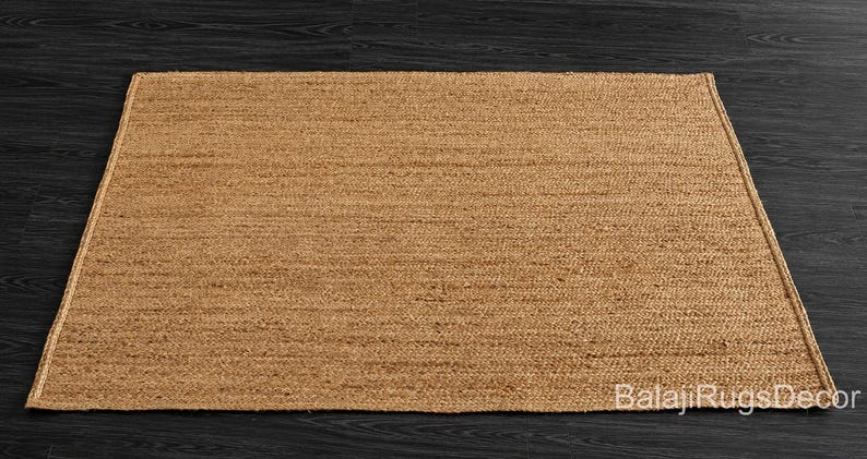 May include: A rectangular, woven jute rug in a natural tan color. The rug has a simple, textured design and is placed on a dark wood surface. The rug's edges are neatly finished, and the overall aesthetic is minimalist and earthy.