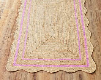 May include: A natural fiber rug with a pink border and a scalloped edge. The rug is made of jute and has a natural, earthy color.