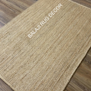 Puede incluir: Una alfombra rectangular de yute tejido en color beige natural. La alfombra tiene una superficie texturizada y está colocada sobre un suelo de madera. Las palabras "BALAJI RUG DECOR" están impresas en blanco en la alfombra.