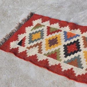 Puede incluir: Una alfombra roja, blanca y marrón con diseños geométricos. La alfombra tiene flecos en un lado.