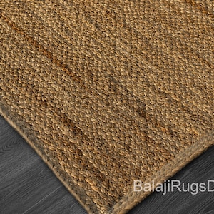 May include: Close-up of a woven jute rug with a natural brown color. The rug has a textured surface and a braided border. The corner of the rug is visible, showing the weave pattern and the dark wood floor beneath. The text "BalajiRugsDecor" is visible.