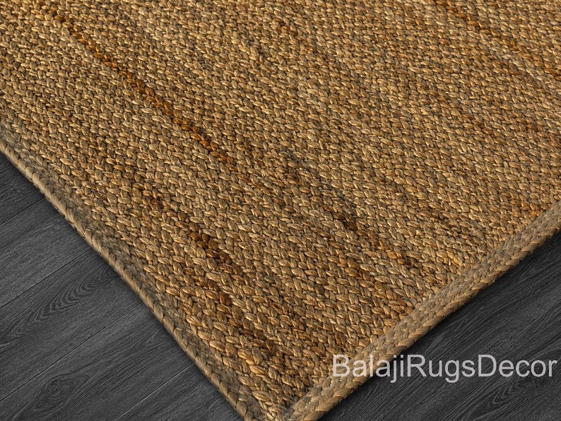 May include: Close-up of a woven jute rug with a natural brown color. The rug has a textured surface and a braided border. The corner of the rug is visible, showing the weave pattern and the dark wood floor beneath. The text "BalajiRugsDecor" is visible.
