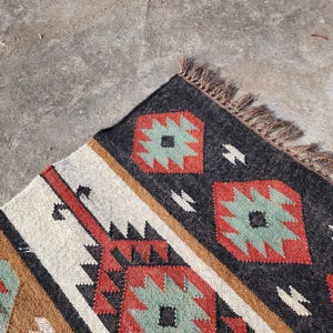 May include: A close-up of a colorful rug with a geometric pattern. The rug is made of wool and has a fringe border. The colors in the rug are black, red, brown, white, and green.