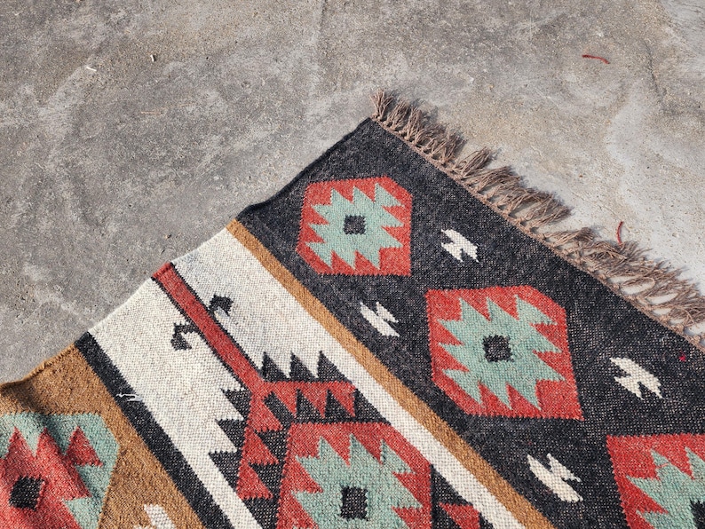 May include: A close-up of a colorful rug with a geometric pattern. The rug is made of wool and has a fringe border. The colors in the rug are black, red, brown, white, and green.