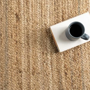 May include: A natural tan braided jute rug with a thick, textured weave. Resting on the rug is a rectangular white book with a dark gray ceramic mug filled with black coffee placed on top of the cover, creating a high-contrast visual arrangement.