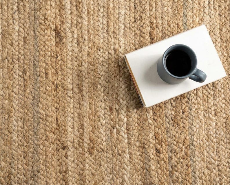 May include: A natural tan braided jute rug with a thick, textured weave. Resting on the rug is a rectangular white book with a dark gray ceramic mug filled with black coffee placed on top of the cover, creating a high-contrast visual arrangement.