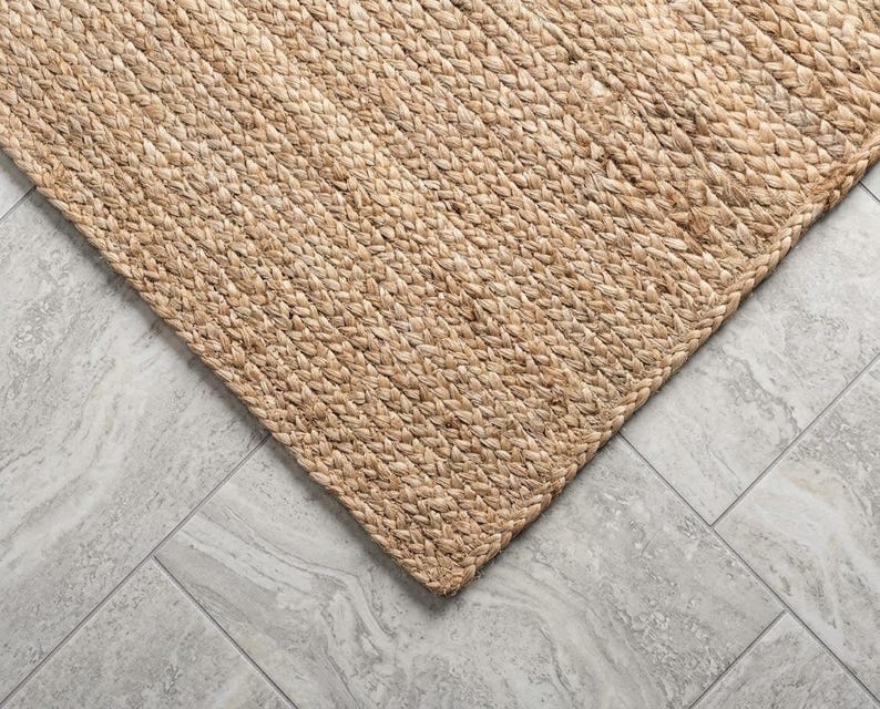 May include: A rectangular area rug crafted from natural tan fibers woven into a thick, braided pattern. The rug features a dense, textured surface with a consistent light brown color throughout, showcasing the intricate interlocking weave design.