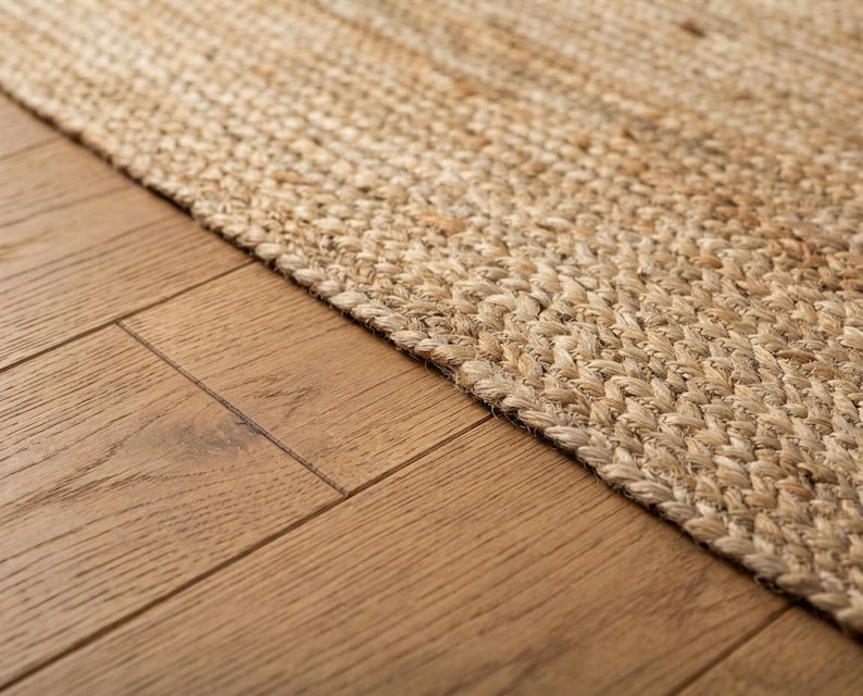 May include: A rectangular area rug crafted from natural tan jute fibers woven in a tight, braided herringbone pattern. The rug features a thick, textured surface with visible variations in the light brown and beige tones of the plant-based material.