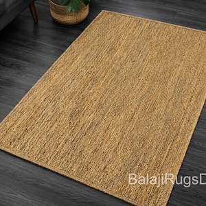 May include: A rectangular, natural-toned jute rug with a woven texture. The rug is placed on a dark wood floor, with a glimpse of a gray sofa and a woven basket with a plant in the background. The rug's surface is a warm, earthy color.