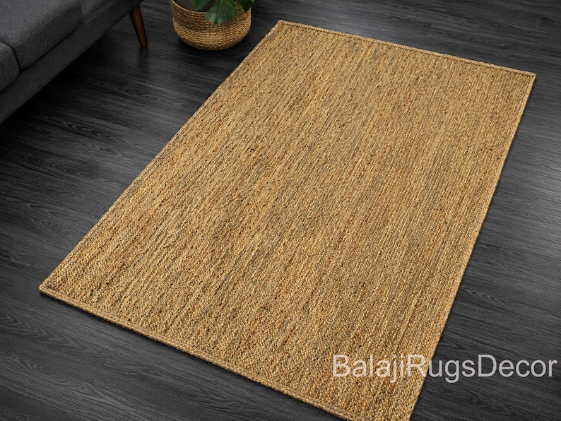 May include: A rectangular, natural-toned jute rug with a woven texture. The rug is placed on a dark wood floor, with a glimpse of a gray sofa and a woven basket with a plant in the background. The rug's surface is a warm, earthy color.