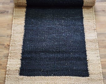 May include: A black and natural jute rug with a black center and a natural jute border. The rug is rolled up and is on a wooden floor.