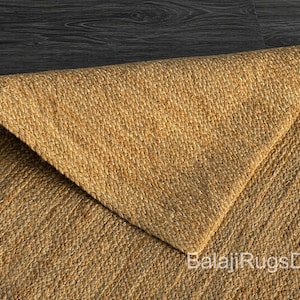 May include: Close-up of a natural jute rug with a woven texture. The rug is a warm tan colour, with a folded corner revealing the underside. The text "BalajiRugsDecor" is visible in the corner.