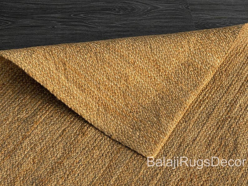May include: Close-up of a natural jute rug with a woven texture. The rug is a warm tan color, with a folded corner revealing the underside. The text "BalajiRugsDecor" is visible in the corner.