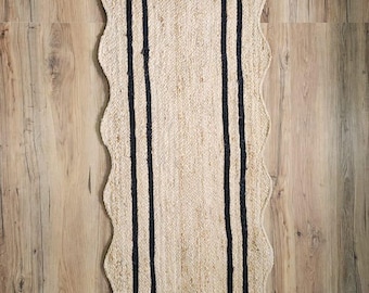 May include: A rectangular jute rug with a scalloped edge and a black border. The rug has a natural beige color and is placed on a wooden floor. The rug's dimensions are approximately 60 inches long and 24 inches wide.