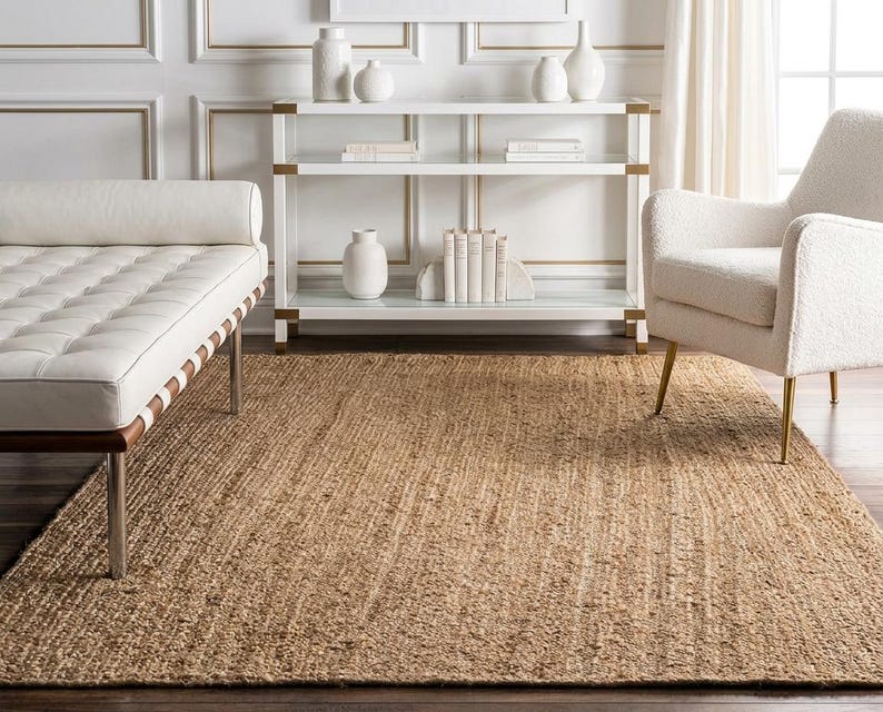 May include: A rectangular area rug made of natural tan woven jute fibers with a coarse, textured surface. The rug is positioned on a dark wooden floor, extending from the foreground toward a white console table, a white daybed, and a white armchair.