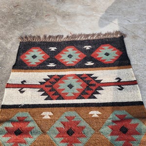 May include: A brown, black, red, and green patterned rug with a fringe border. The rug has a geometric design with repeating diamond shapes.