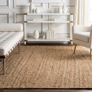 May include: A rectangular area rug made of natural tan woven jute fibres with a coarse, textured surface. The rug is positioned on a dark wooden floor, extending from the foreground towards a white console table, a white daybed, and a white armchair.