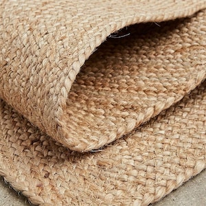 May include: A tan-colored rug made of woven natural fibers with a thick, textured, braided construction. The material features a visible cross-hatch pattern with a folded corner that reveals the dense, layered structure of the floor covering.