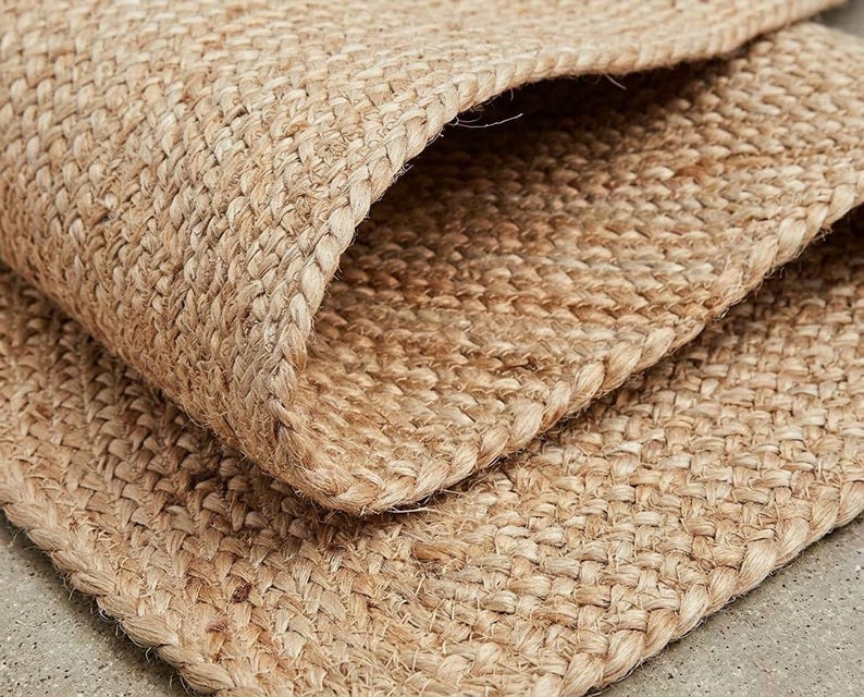 May include: A tan-colored rug made of woven natural fibers with a thick, textured, braided construction. The material features a visible cross-hatch pattern with a folded corner that reveals the dense, layered structure of the floor covering.