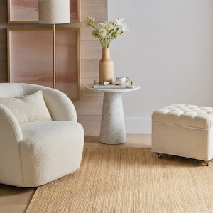 May include: A white boucl&eacute; swivel armchair with a matching lumbar pillow, a white marble pedestal side table, and a square cream-colored tufted ottoman with small wheels. The furniture is arranged on a natural fiber woven area rug.