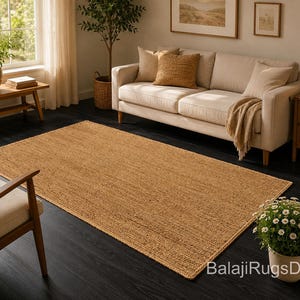 May include: A rectangular, woven jute rug in a warm tan color, displayed on a dark wood floor. The rug is placed in a living room setting, with a cream-colored sofa, wooden furniture, and natural light.