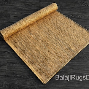 May include: A rolled-up, rectangular jute rug in shades of tan and brown. The rug is partially unrolled, revealing its woven texture. The background is a dark wood surface. The text "BalajiRugsDecor" is visible.