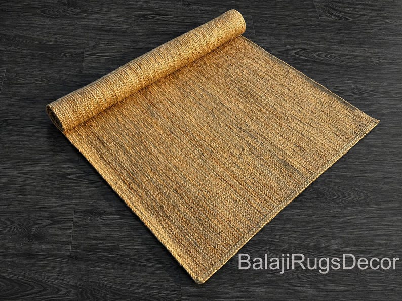 May include: A rolled-up, rectangular jute rug in shades of tan and brown. The rug is partially unrolled, revealing its woven texture. The background is a dark wood surface. The text "BalajiRugsDecor" is visible.
