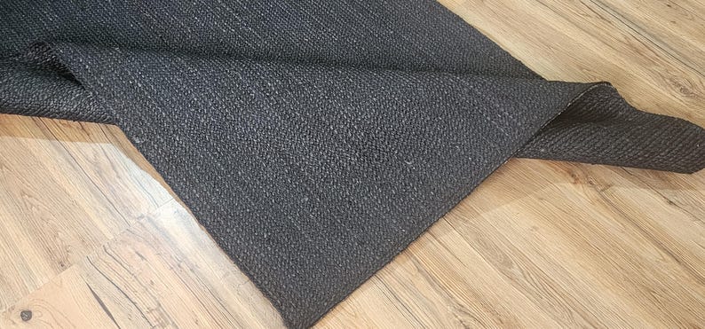 May include: A black woven rug with a textured surface. The rug is folded in half and lying on a wooden floor.