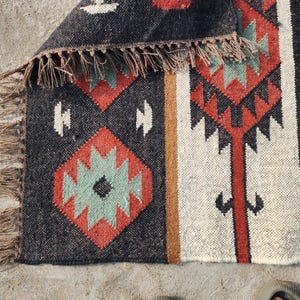 May include: A black, white, red, and green patterned rug with a fringe border. The rug has a geometric design.