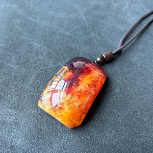 May include: A rectangular amber pendant with a brown cord necklace. The amber is a mix of orange, yellow, and red colors with a cloudy, translucent appearance.